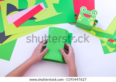 The kid makes a frog out of paper. Princess frog is green with a crown. Children's master class. School and kindergarten.