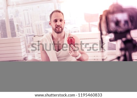 Healthy lifestyle. Full length of enthusiastic handsome bearded sportsman holding red pepper in hand and sitting on the floor while talking about useful diet in front of camera