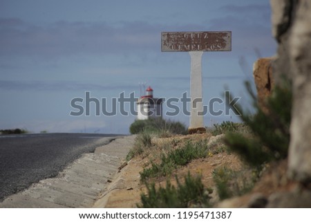 A worn out concrete sign next to a road, with a white lighthouse in the background on a sunny day in Peniche, Portugal.