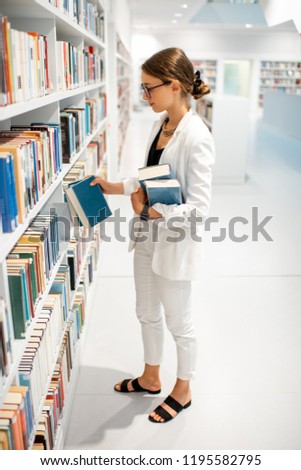 Young businesswoman searching books standing near the bookshelves at the modern white library
