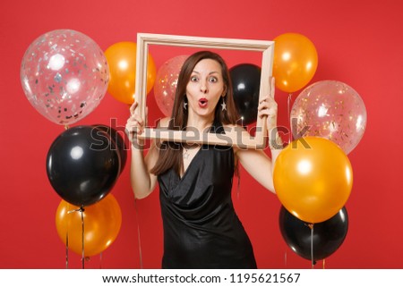 Excited young woman in little black dress celebrating and holding picture frame on bright red background air balloons. International Women's Day, Happy New Year, birthday mockup holiday party concept