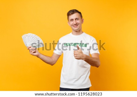 Man in white t-shirt with written inscription green title volunteer hold lots dollars banknotes, cash money isolated on yellow background. Voluntary free assistance help, charity grace work concept