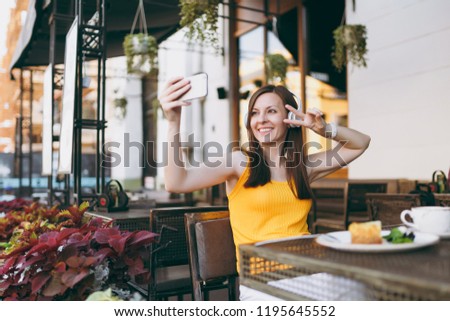 Attractive smiling woman in outdoors street coffee shop cafe sitting at table, listen music in headphones, doing selfie shot on mobile phone, relaxing in restaurant free time. Lifestyle rest concept