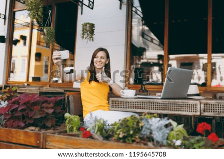 Happy smiling girl in outdoors street coffee shop cafe sitting at table with laptop computer doing taking selfie shot on mobile phone in restaurant during free time. Mobile office freelance concept