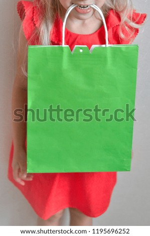 A little girl holds package in teeth enjoys shopping and sale on a light background. Child in good mood