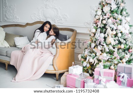 Young mother sits on sofa with plaid and kisses her little daughter against background of Christmas tree and decorations.