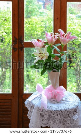 Pink and white lilies in a ceramic vase, tied with a pink ribbon on a white lace on a wooden table, attached to a glass door and a droplet attached to make it fresh.