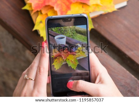 The girl on the phone makes a photo of yellow autumn leaves on the book a mug of coffee