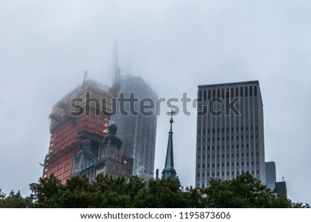 A modern building stands next to a historical building with a tall speeple. A tall skyscraper is being constructed behind the  historic building. A crane is on the tall building. Low clouds obscure.