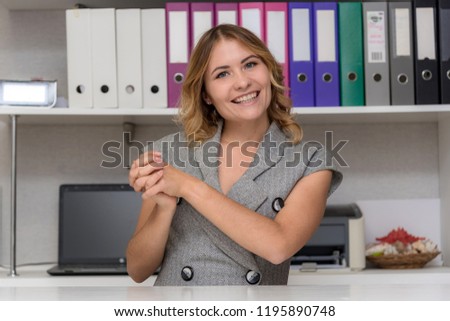 Portrait of a beautiful blonde girl in the office at the table. She is sitting right in front of the camera, smiling and looking happy.