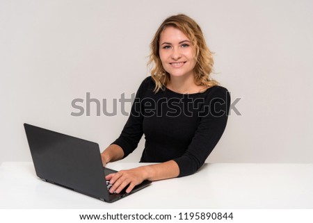 Portrait of a beautiful blonde girl on a white background at the table with a laptop. She is sitting right in front of the camera, smiling and looking happy.