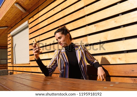 Young brunette woman in multi-colored jacket sitting at a wooden table looking at mobile phone
