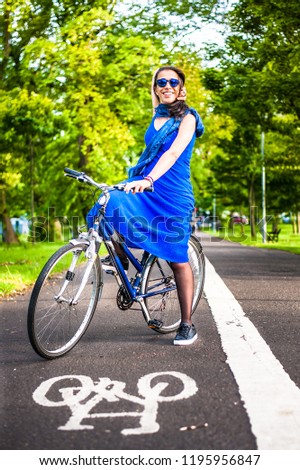 Young brunette girl in blue dress and sunglasses sitting on a bicycle stopped at a bicycle sign on asfalt road.