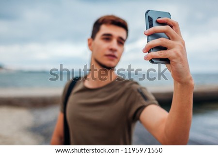 Young man taking selfie using phone on cloudy beach. Handsome sportive guy walking by pier