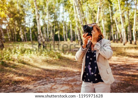 Middle-aged woman taking pictures using camera in autumn forest. Senior woman walking and enjoying hobby