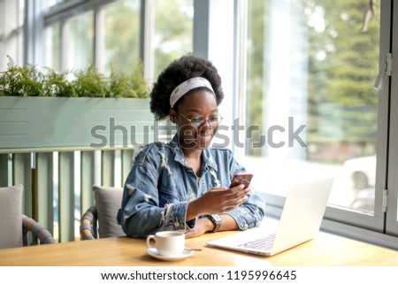 Young dark-skinned business woman in denim shirt sitting at table in cafe, talking on smart phone while looking on screen of laptop standing on table near the cup of coffee. Freelancer working.