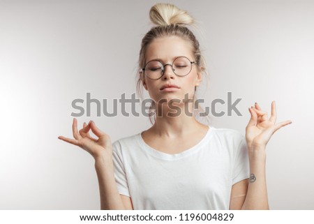 Concentrated beautiful young woman with blonde hair knot dressed in white t-shirt keeping eyes closed while meditating indoors, practicing peace of mind, keeping fingers in mudra gesture