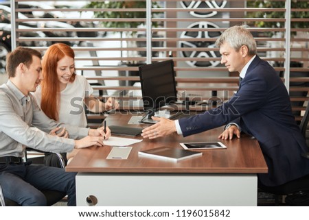 Couple of buisness people listening to conditions of rental agreement for signing, sitting at table with the agent at car rental salon