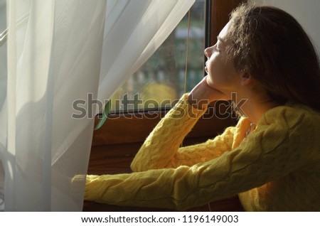 Portrait of a beautiful girl who looks out the window sitting in a yellow sweater at home, horizontal photo