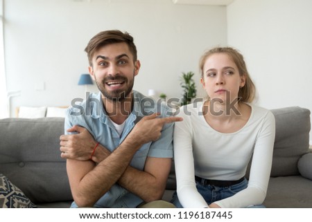 Millennial married couple sitting together on couch in living room at home. Boyfriend and girlfriend having video call funny male pointing on dissatisfied female complains talking with friends Royalty-Free Stock Photo #1196187445