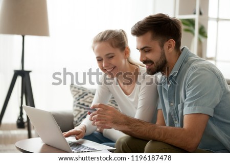 Happy married couple sitting in living room on couch at home, using computer purchasing online. Husband holding credit card customers making secure payment via internet. Technology, e-banking concept Royalty-Free Stock Photo #1196187478