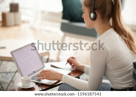 Student girl sitting at home looking at laptop screen listening exercise, studying foreign languages online making notes, side view. Call center operator working indoors. Technology e-learning concept Royalty-Free Stock Photo #1196187541