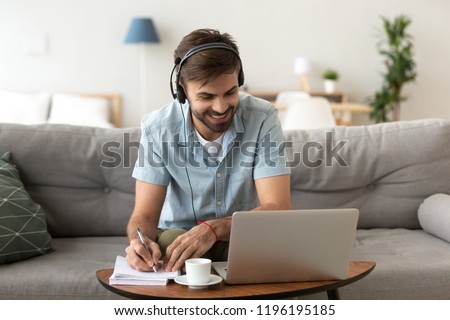 Man sitting on couch in living room at home enjoying studying using laptop and headset looking at device screen listening audio making some notes. Male has lesson online e-learning in internet concept Royalty-Free Stock Photo #1196195185