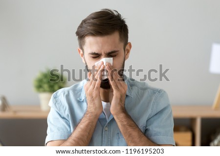 Young sick man sitting at office room sneeze holding tissue handkerchief blowing wiping his running nose. Headshot Male feels unwell caught influenza cold seasonal infection or having allergy concept Royalty-Free Stock Photo #1196195203