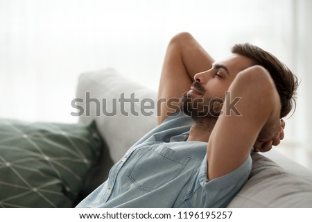 Close up side view serene man smiling sitting on couch at home. Male has a break after work or study closing eyes putting hands behind head relaxing thinking, feels happy breathing fresh air concept Royalty-Free Stock Photo #1196195257