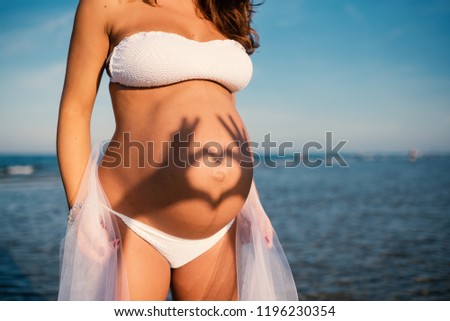 Young pregnant woman portrait on the beach in the summer with heart shape shadow. Real lifestyle image.