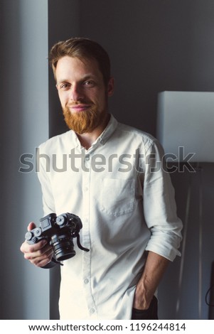 Portrait of young hipster man with photo camera in hands. Freelance photographer with camera looking in camera. Male photographer with beard in white short. Man photographer in white shirt with camera