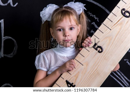Cute schoolgirl standing before the chalkboard as a background holding the huge ruler diagonally. Enlarged view.
