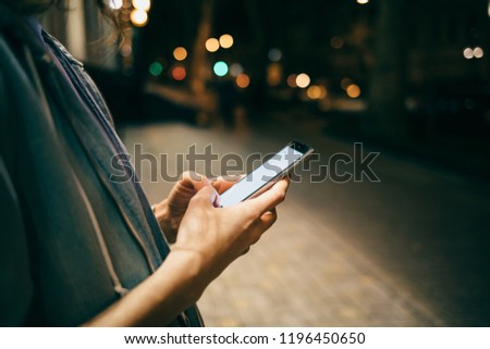 Girl typing message on smart phone on background bokeh lights in night city. Close-up young woman online in the dark time on illuminated street outside.