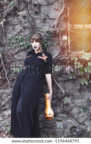 Young brunette woman holding pumpking, posing outside in black halloween outfit.