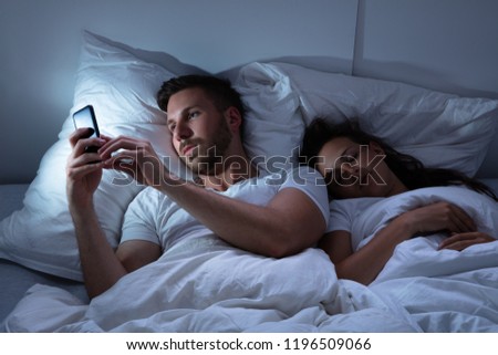 Young Man Using Cellphone While Her Wife Sitting On Bed At Night Royalty-Free Stock Photo #1196509066