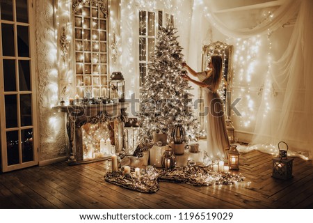 beautiful blonde woman in a white long dress  is standing in a beautiful bright room with decoration near shiny Christmas tree and fireplace with garlands lanterns, candles, gifts at night