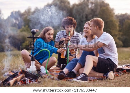 Teenagers have fun. Joyful girls and boys spend weekend outdoor on picnic, smoke hookah, clink bottles with energetic drinks, celebrate starting holidays. Friendship, youth and lifestyle concept