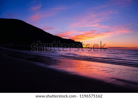 Blue, pink and orange sunset reflected on the water and waves, with the black mountain and beach shore of Mont-Louis, Gaspésie, QC, Canada
