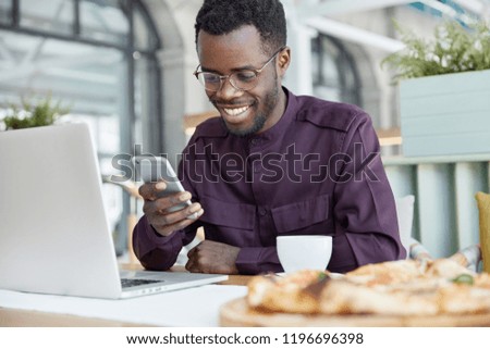 Cheerful dark skinned guy installs app on laptop computer, shares media on smart phone, uses wireless internet connection in cafeteria, searches website, drinks hot beverage, has shining smile