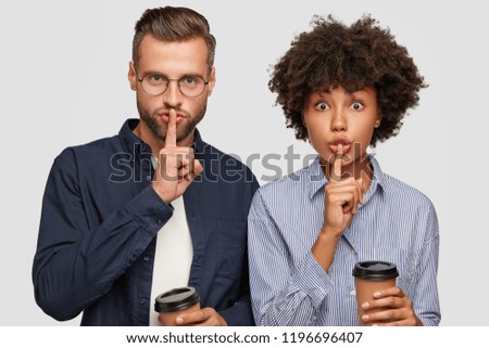 Horizontal shot of serious mixed race female and male keep index fingers over mouth, make shush gesture, hold disposable cups of coffee, isolated over white background. Shush, be silent please