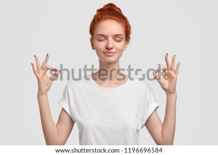 People, yoga and mediatation concept. Relaxed foxy girl with freckled soft skin enjoys peaceful atmosphere, keeps hands in mudra sign, relaxed after intense day, isolated over white background