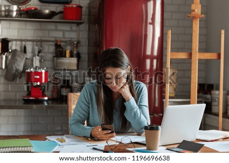 Female emloyee with dark long hair, dressed in stylish shirt, uses mobile phone for searching internet, works on business report, drinks takeaway coffee, uses laptop computer, chats with client