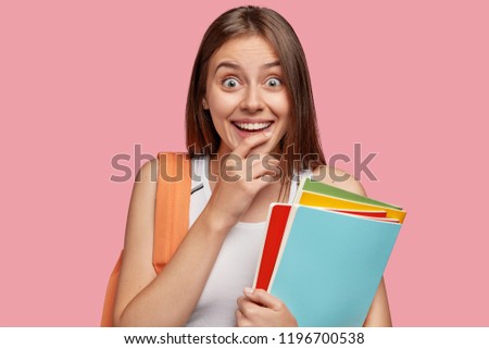 Indoor shot of pleasant looking cheerful woman with toothy smile, keeps hand on chin, looks in amazement, dressed in casual outfit, carries books, going to school, has rucksack on back, models indoor