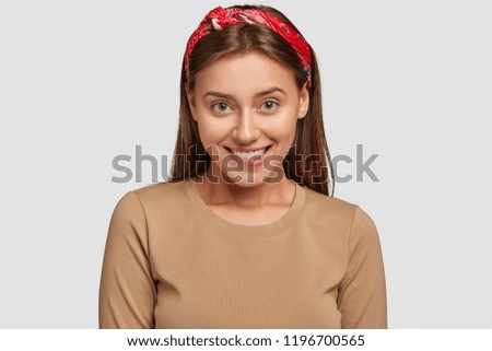 Photo of pleased young female model with toothy expression, looks joyfully into camera, wears red bandana, has friendly talk with guy, isolated over white background. Emotions and facial expressions