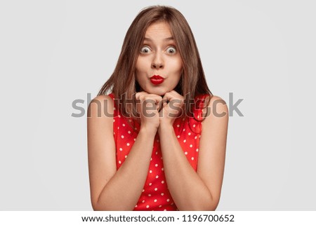 Surprised brunette young woman with bugged eyes, keeps red lips pressed and rounded, holds chin, stares at camera, notices something amazing, has long dark hair, poses against white background
