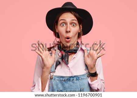 Oh my God! Terrified beautiful Caucasian woman gasps from fear, holds palms near chest, being astonished with horrible event on farm, dressed in black headgear and denim overalls, isolated on pink