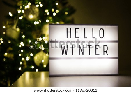 winter and christmas concept - lihtbox with text hello winter in dark room with decorated christmas tree