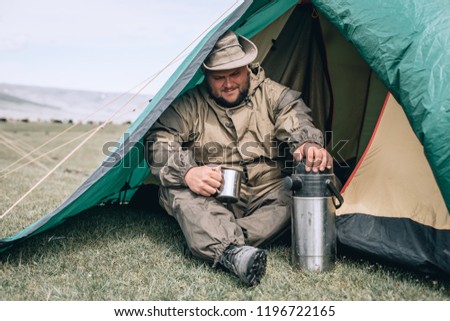 Young man in hat, drinking tea or coffee in mountains. Mountain camping concept. Traveler man with beard sit near the tent neat the mountain lake . Man traveler hands holding cup of tea. 