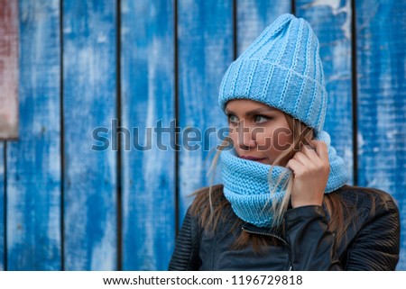 Portrait of a beautiful young model in blue knitted hat standing near blue wall on an autumn day. Autumn warm photo.Woman smiling and look at the camera, joyful cheerful mood.