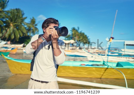 Photography and travel. Young man with rucksack taking photo with his camera on the sea beach near fishing boats.